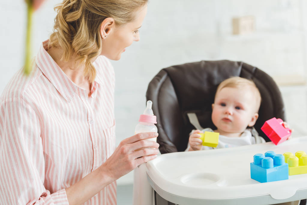 young mother holding feeding bottle infant daughter sitting baby chair